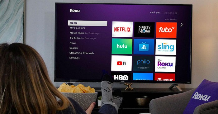 A woman sitting on a couch in front a TV displaying the Roku homescreen.