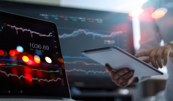 An analyst using a tablet computer with computer screens displaying stock charts in the background.